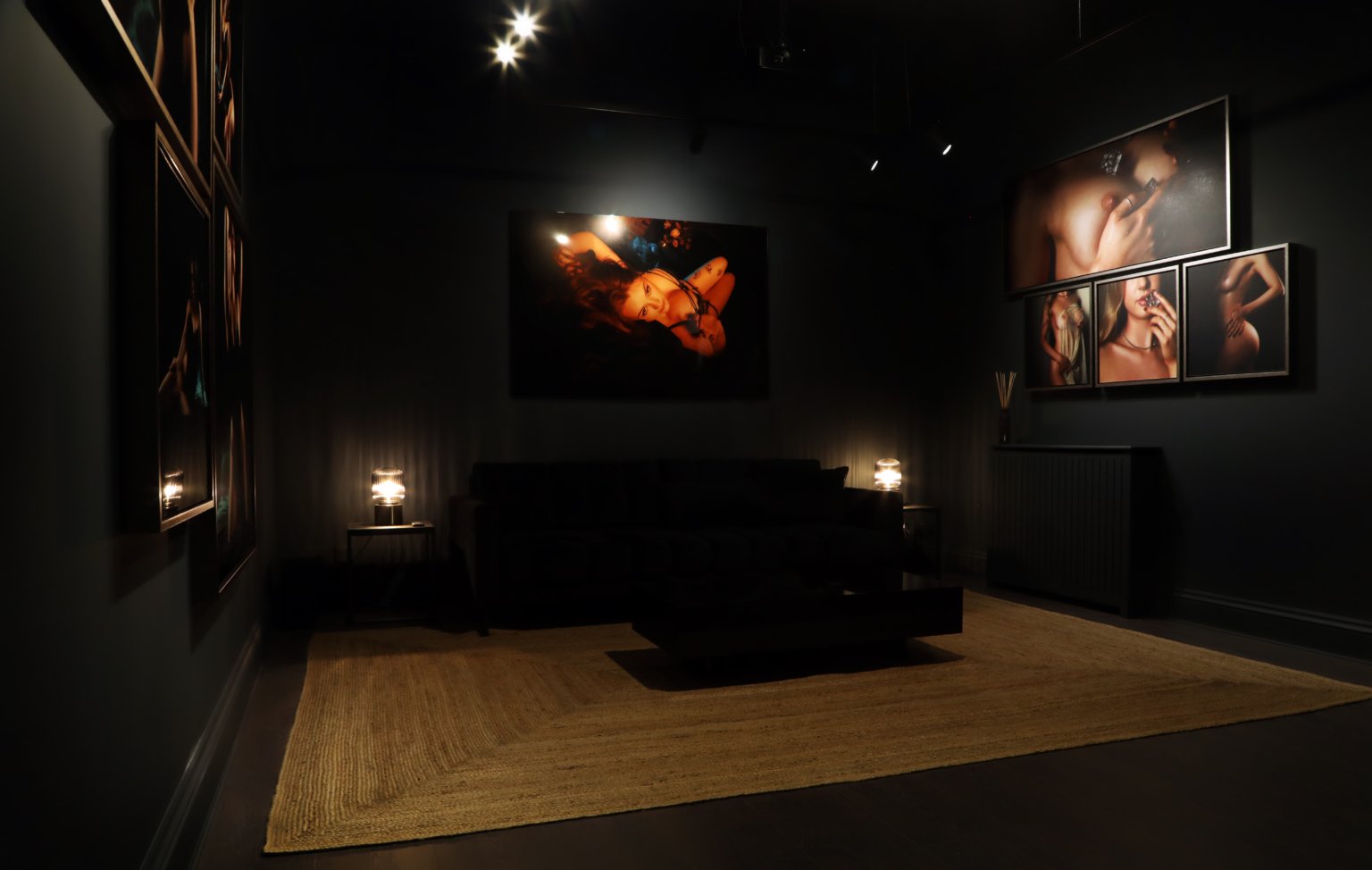 dark room with boudoir wall art mounted on the walls, two lamps and a black couch